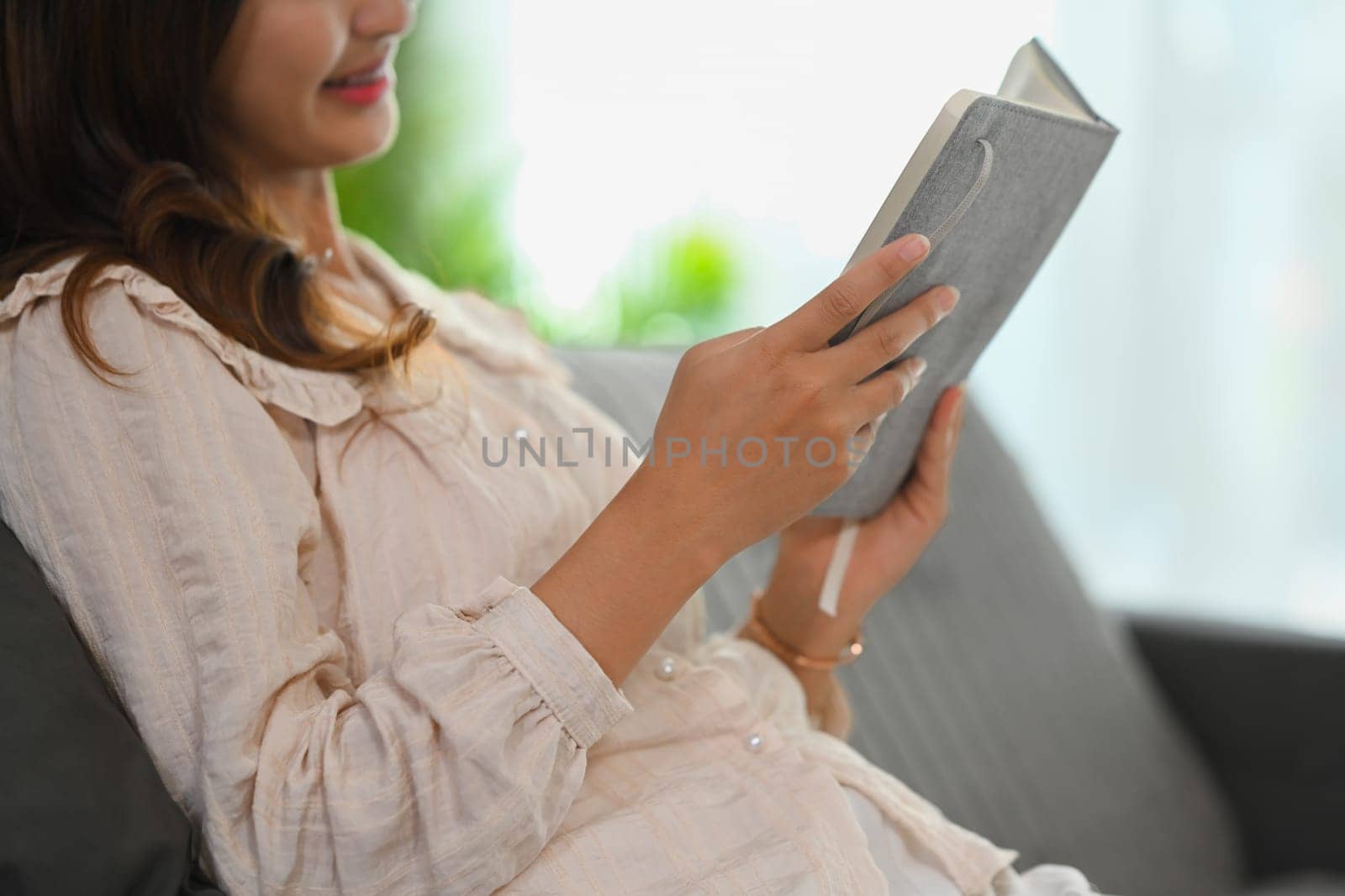 Cropped shot happy young woman in casual clothes reading book on sofa at home by prathanchorruangsak