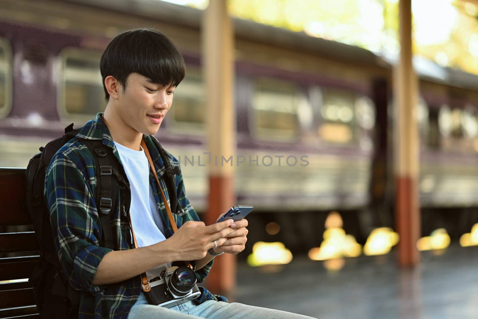 Young man with a backpack sitting at the train station and using mobile phone.