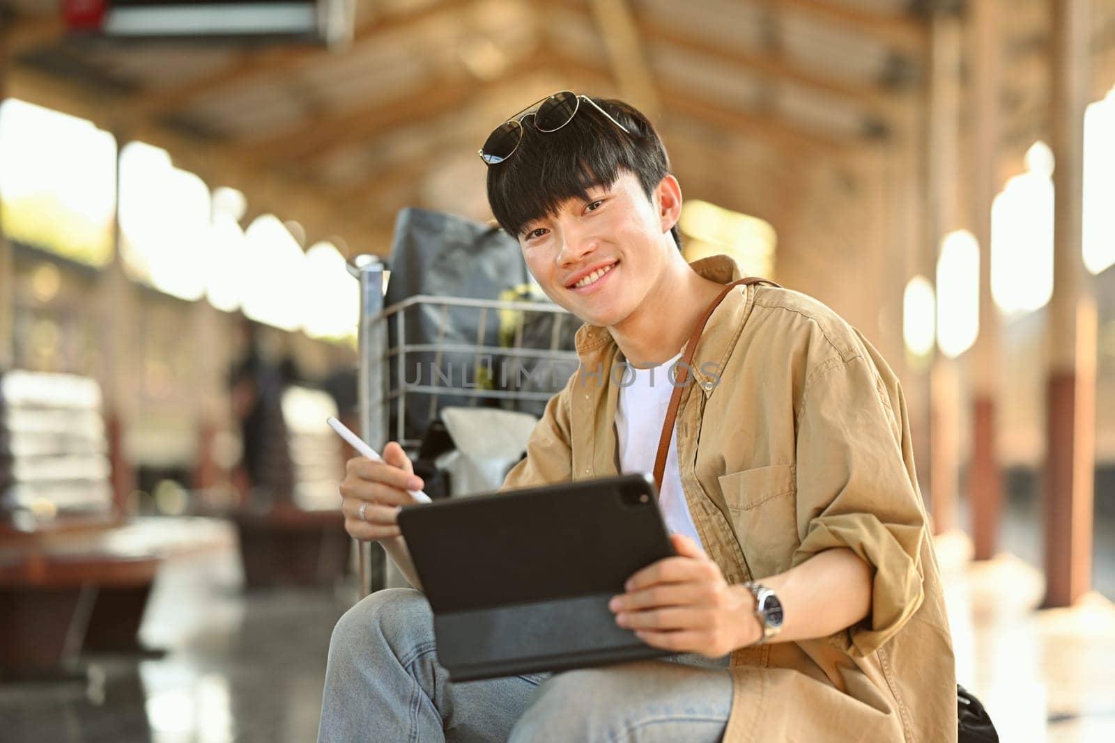 Portrait of smiling Asian man tourist using digital tablet while waiting train at railway station.