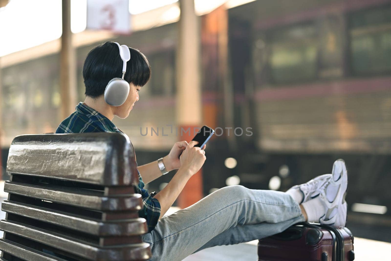Young man in headphone using mobile phone during waiting for train at railway station.