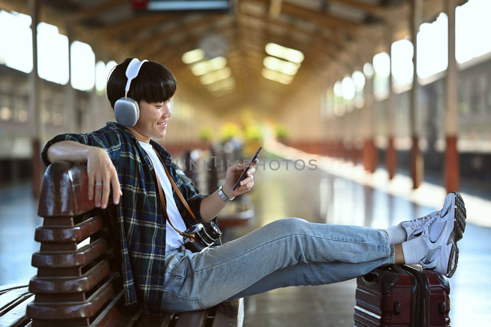 Happy young man in headphone using mobile phone during waiting for train at railway station.