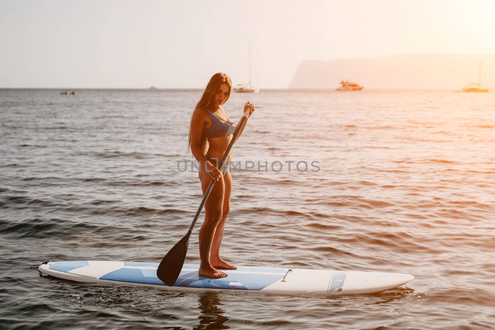 Woman sea sup. Close up portrait of happy young caucasian woman with long hair looking at camera and smiling. Cute woman portrait in a blue bikini posing on sup board in the sea by panophotograph