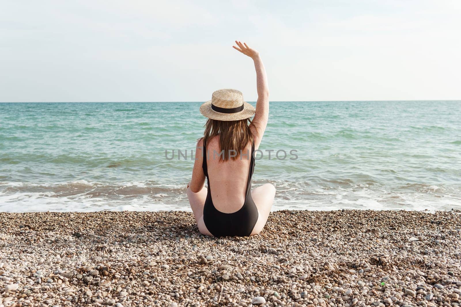 Woman in black swimsuit and straw hat on beach, seaside. View from the back. Summer vacation at the sea