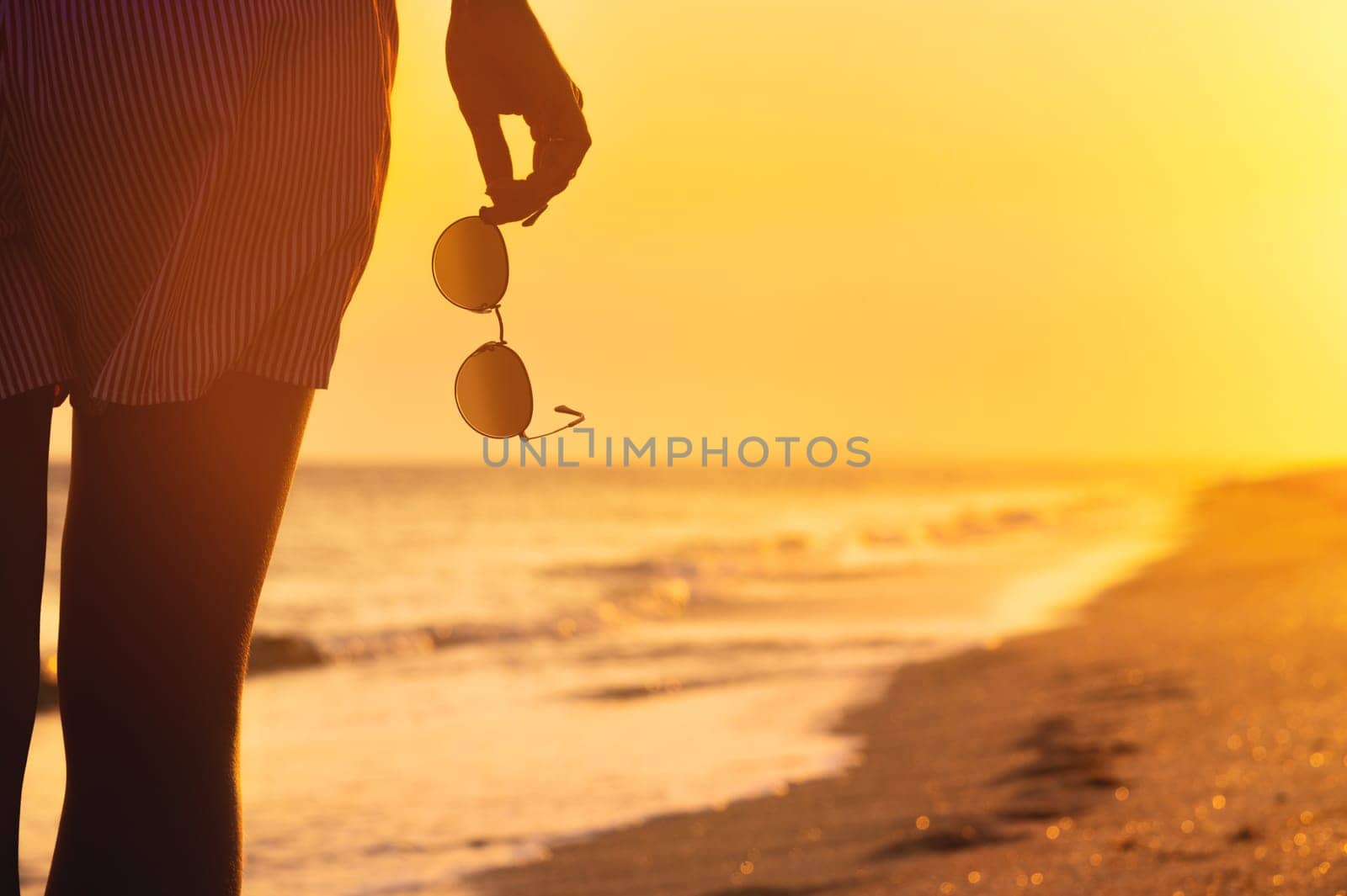 woman in shorts stands on the shore with sunglasses in her hand, close-up. Warm colors, beach holiday at sunset by yanik88