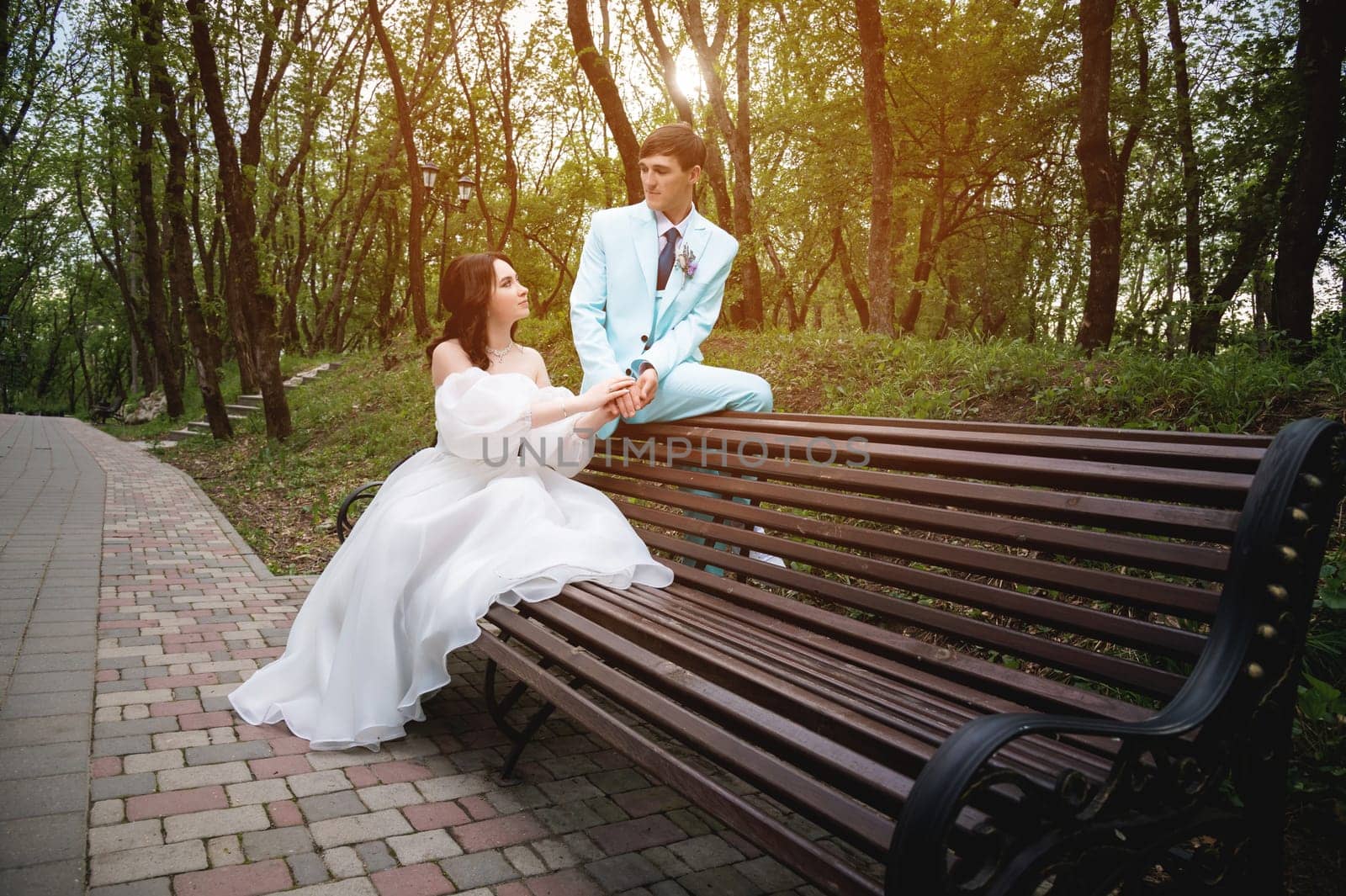 European bride and groom are sitting on a park bench. Newlyweds admiring each other while walking through a green summer park by yanik88
