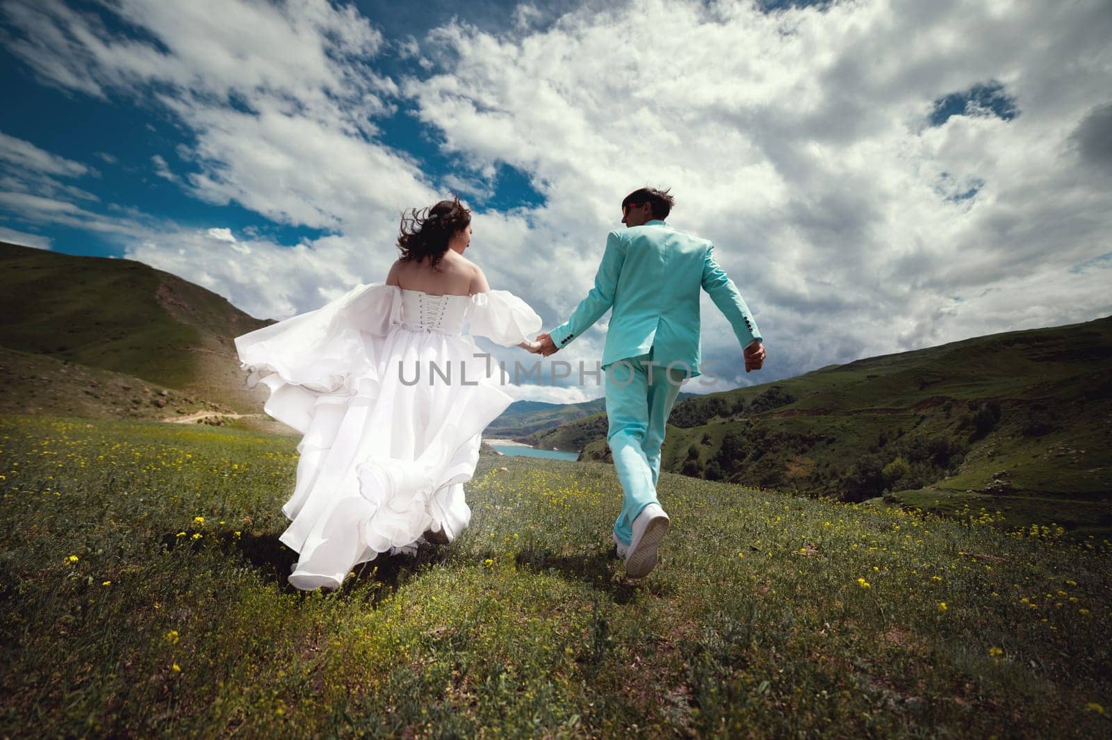 Rear view of running loving newlyweds holding hands, a bride in a wedding dress and a wife in a turquoise suit running across a field against the backdrop of green hills.