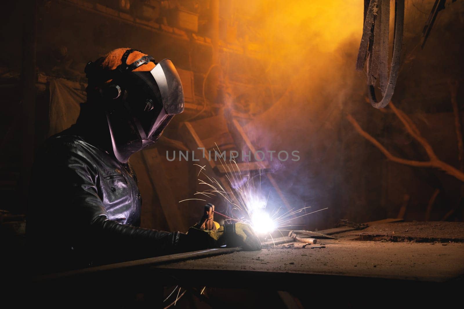 A welder in a workshop welds a metal part. General plan of an old cluttered garage where a man in protective gloves and a mask is making.