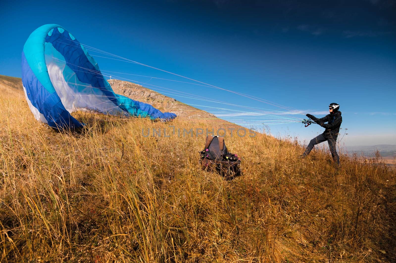 The paraglider is preparing to take off. A colorful kite lies on the grass, in the background there is a beautiful landscape, clear blue sky.