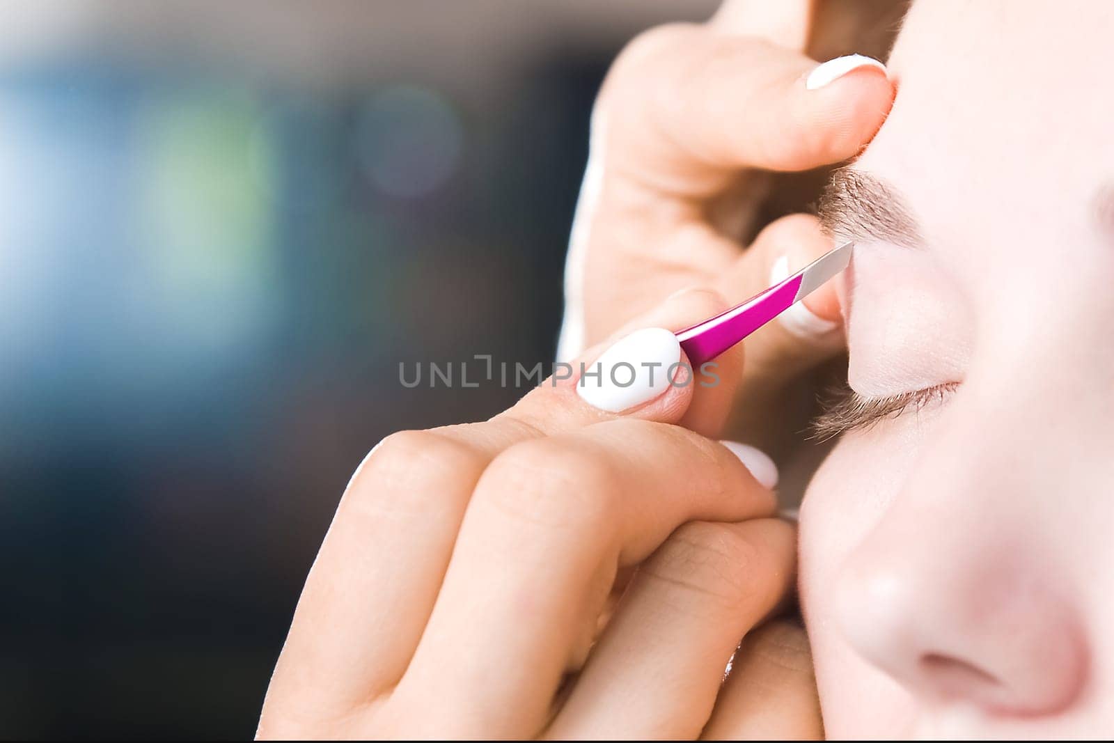 Extreme close-up of a young woman plucking eyebrow hair with tweezers in a beauty salon.