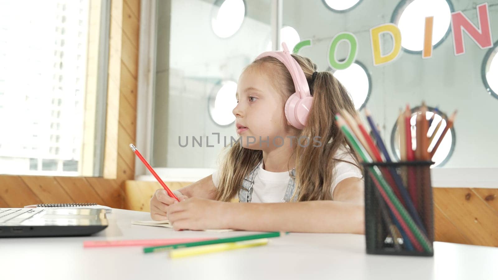 Cute smart girl doing classwork or online learning while listen to teacher. Happy student writing, drawing, working on paper while wearing headphone with laptop and colorful color pencils. Erudition.