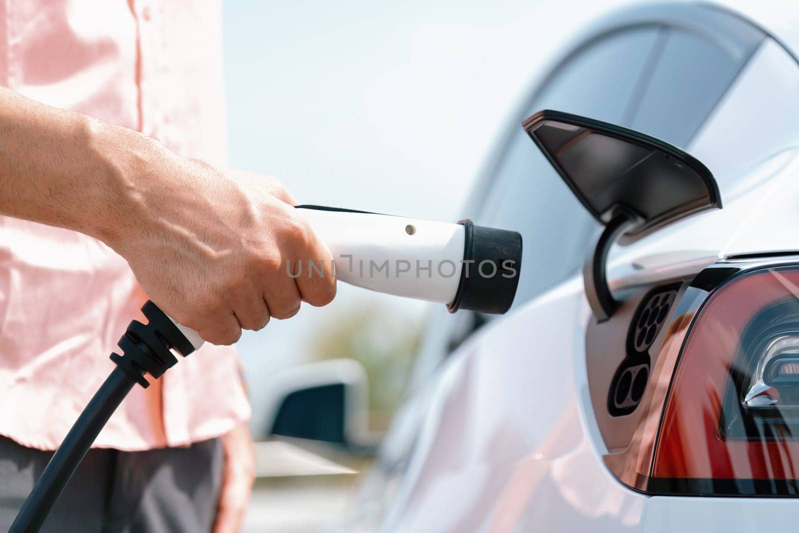 Hand insert EV charger plug into electric vehicle to recharge EV car battery from outdoor charging station. Fresh daylight environment with alternative clean and sustainable energy concept. Perpetual