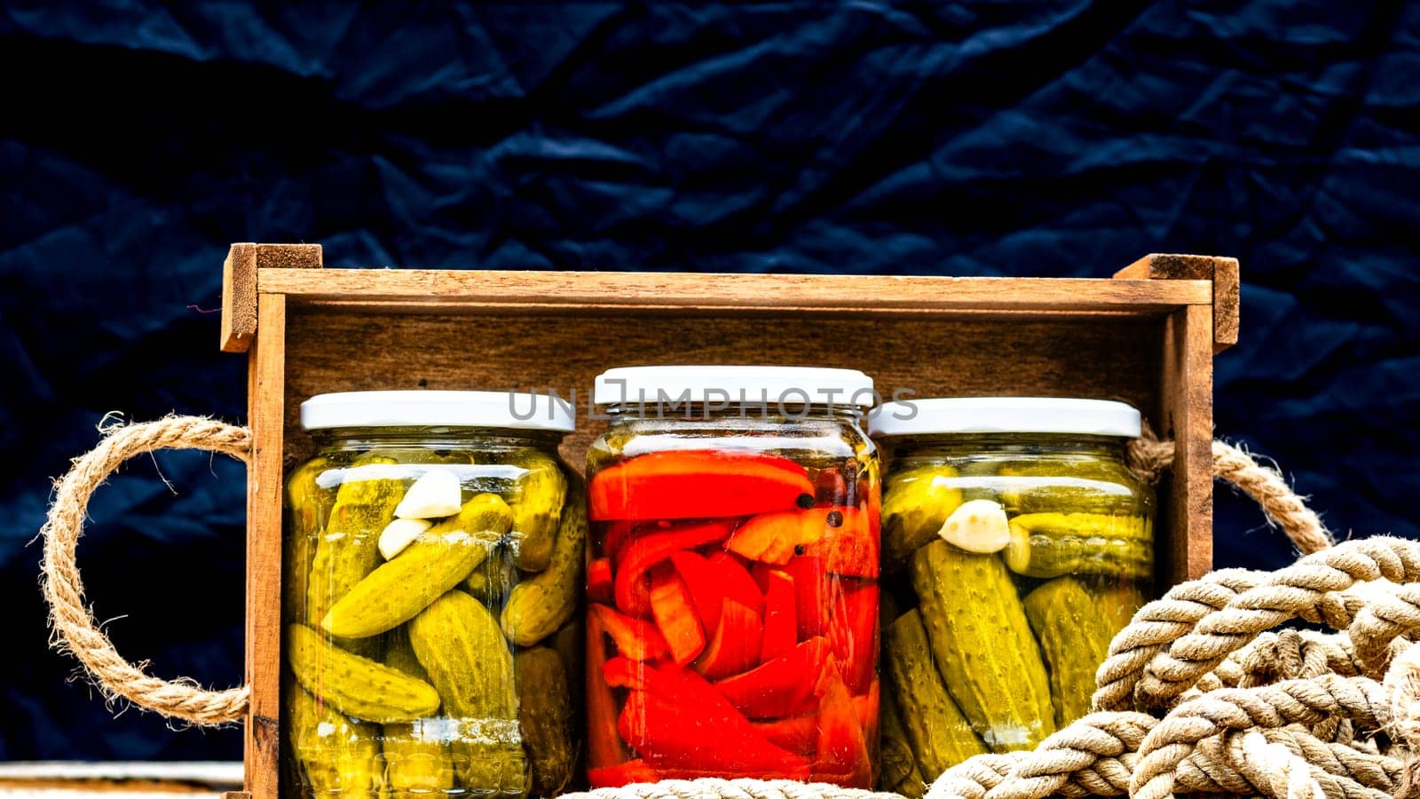 Wooden crate with glass jars with pickled red bell peppers and pickled cucumbers (pickles) isolated. Jars with variety of pickled vegetables. Preserved food concept in a rustic composition.