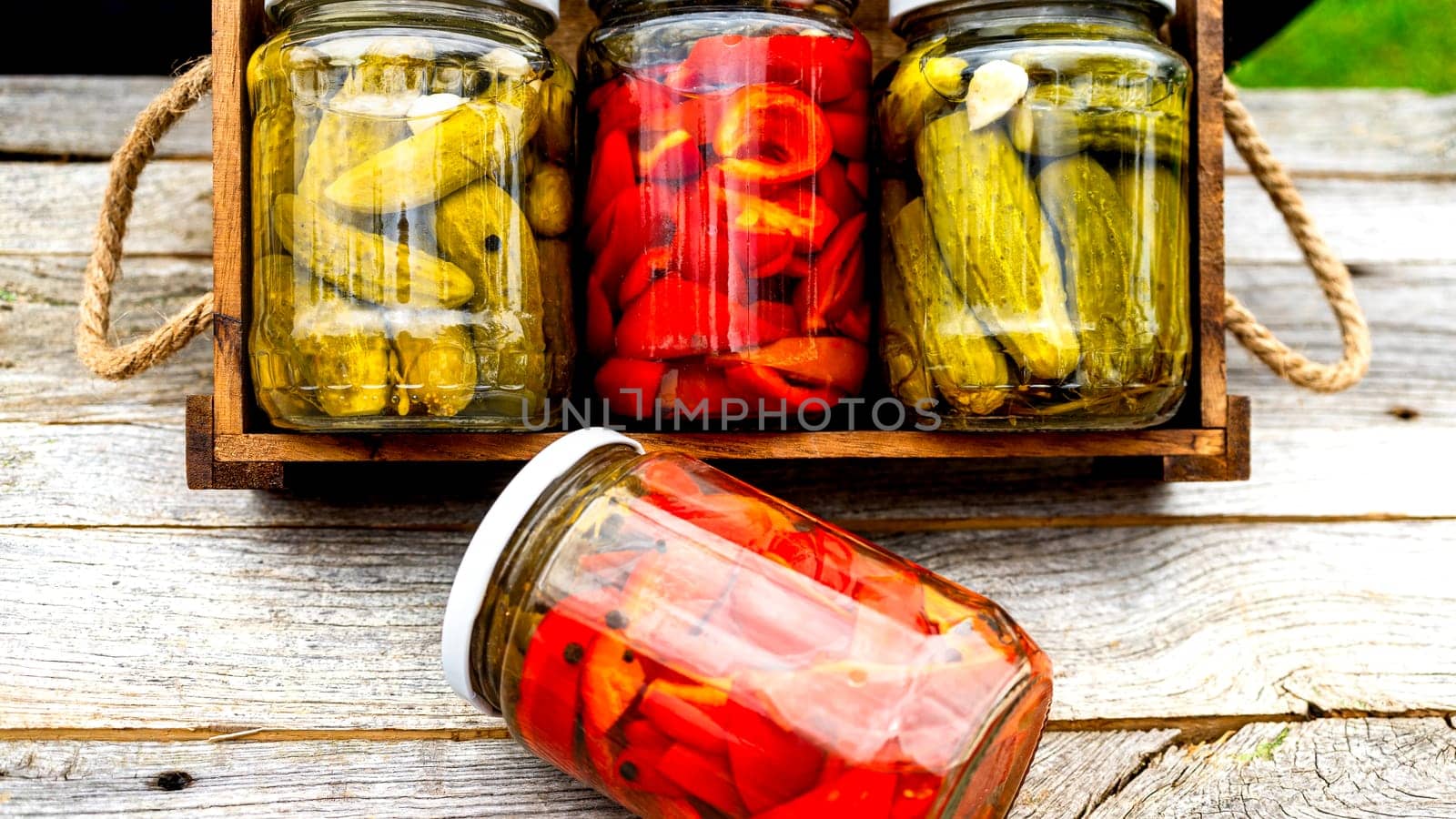 Glass jars with pickled red bell peppers and pickled cucumbers (pickles) isolated in wooden crate. Jars with variety of pickled vegetables. Preserved food concept in a rustic composition. by vladispas