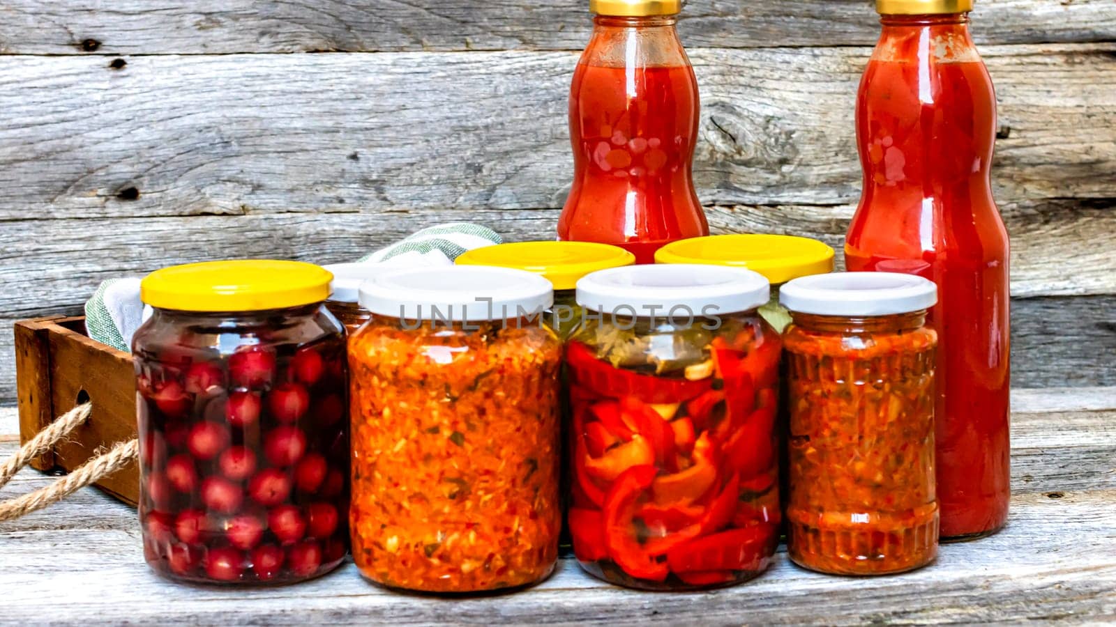 Jars with variety of canned vegetables and fruits, jars with zacusca and bottles with tomatoes sauce. Preserved food concept in a rustic composition.