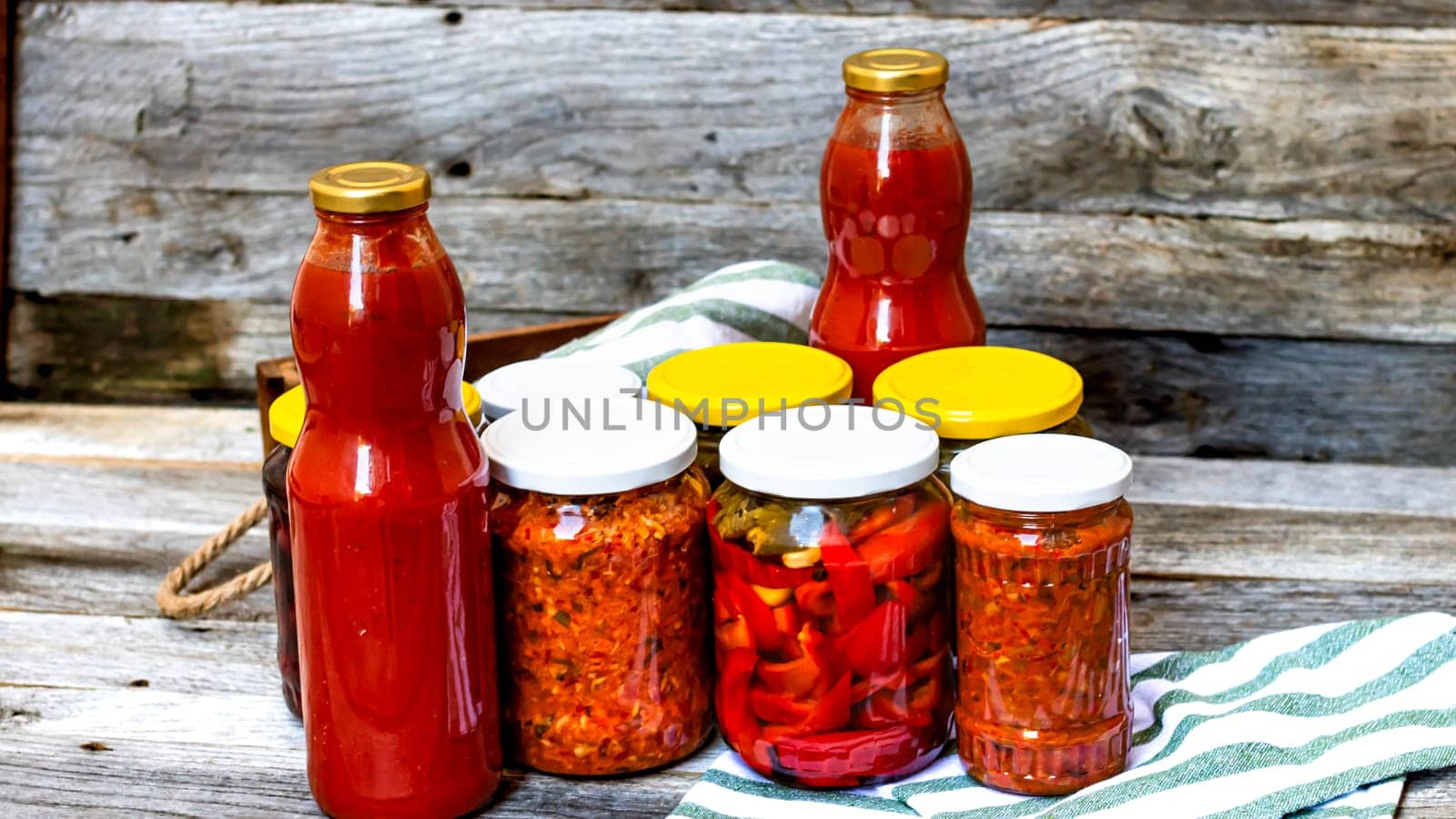 Jars with variety of canned vegetables and fruits, jars with zacusca and bottles with tomatoes sauce. Preserved food concept in a rustic composition. by vladispas