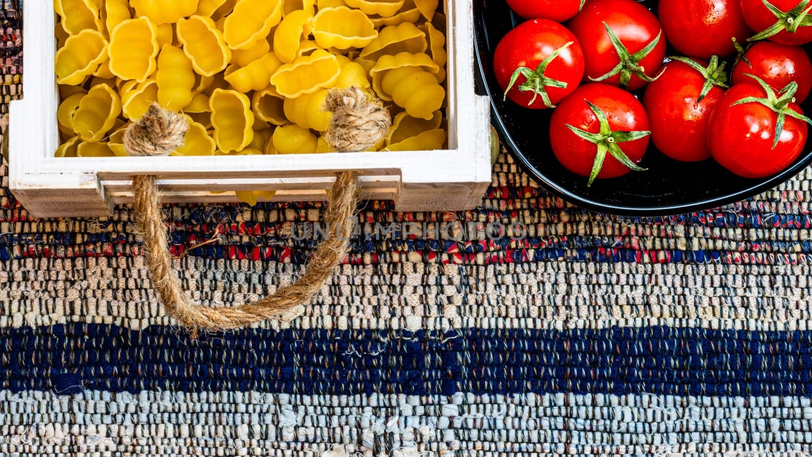 Top view of pasta in a wooden crate and fresh ripe cherry tomatoes in a black bowl. Ingredients and food concept