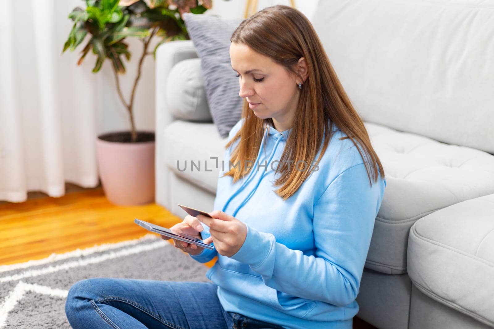Young woman in a blue hoodie focused on browsing her smartphone while seated on the floor at home