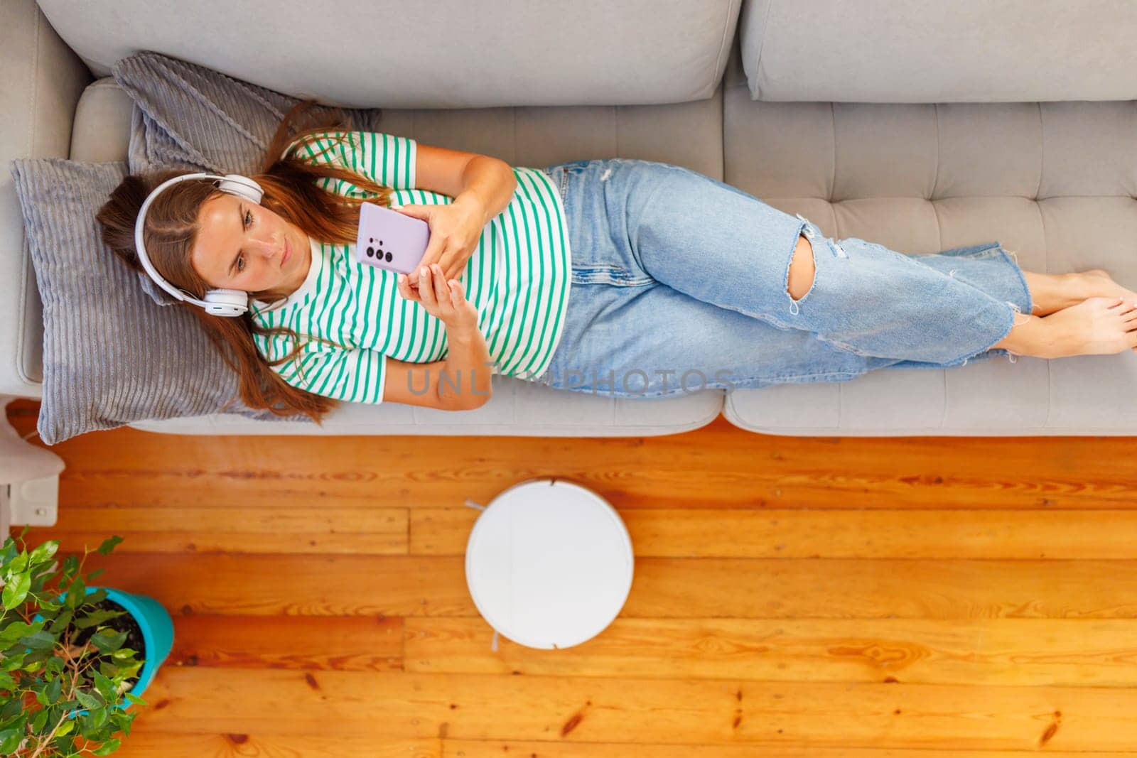 A young woman enjoys a peaceful moment on a sofa, listening to music on her headphones while holding a smartphone, with a robotic vacuum cleaner in the background.