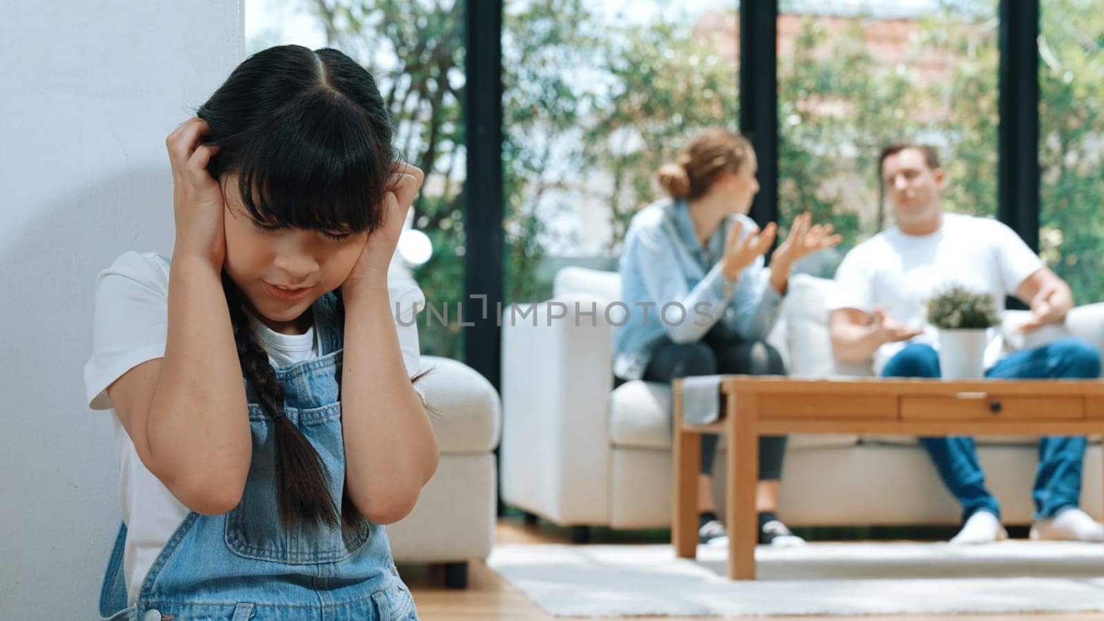 Stressed and unhappy young girl huddle in corner, cover her ears blocking sound of her parent arguing in background. Domestic violence at home and traumatic childhood develop to depression. Synchronos