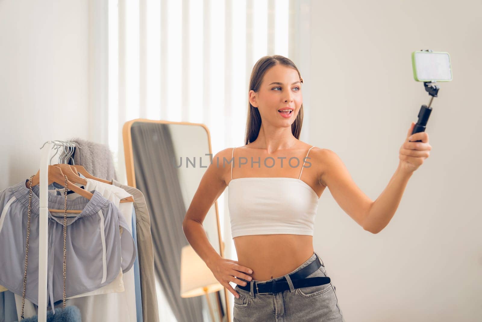 Young woman making fashion video content for social media. Blogger smiles to camera using selfie stick and light ring while making persuasive online clothing sell to audience or followers. Unveiling