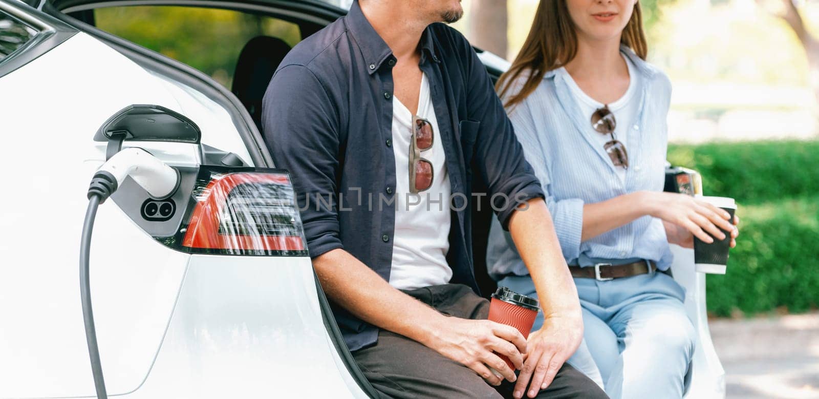 Lovely young couple drinking coffee while recharging battery for electric car during road trip travel EV car in natural forest or national park. Eco friendly travel during vacation and holiday. Exalt