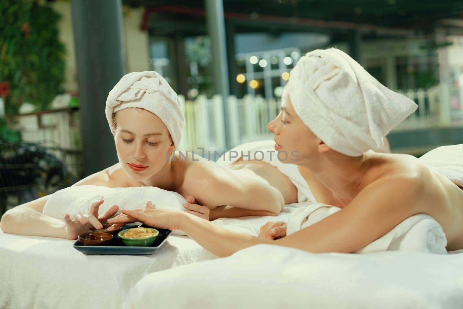 Couple of beautiful young girls lie on spa bed during interested in homemade beauty facial mask. Attractive woman touching herbal facial mask. Surrounded with nature environment. Tranquility.