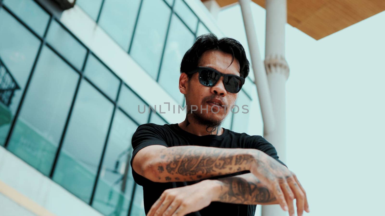 Hispanic dancer looking at camera while dancing at sky scrapper. Attractive handsome hipster moving his hands to hip hop music at modern mall with low angle camera. Outdoor sport 2024. Endeavor.