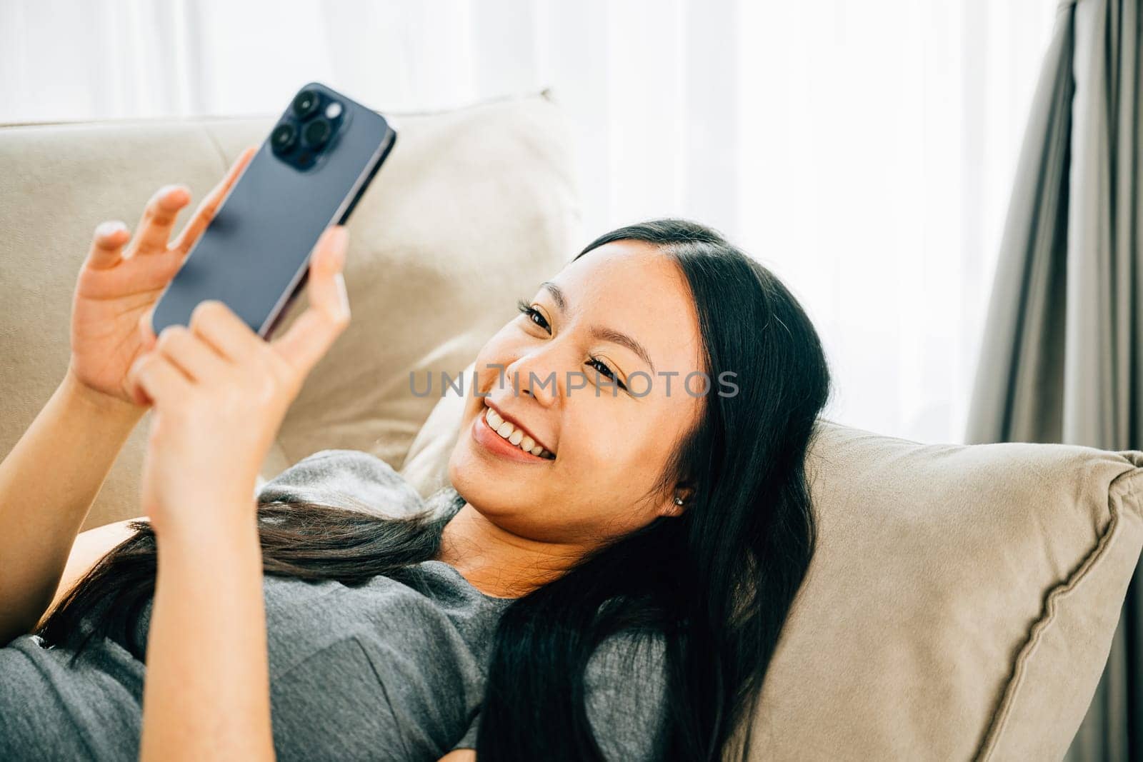 Happy woman on comfortable sofa texts on smartphone smiling. Enjoying relaxation chatting and shopping online. Modern technology for communication and connection at home.