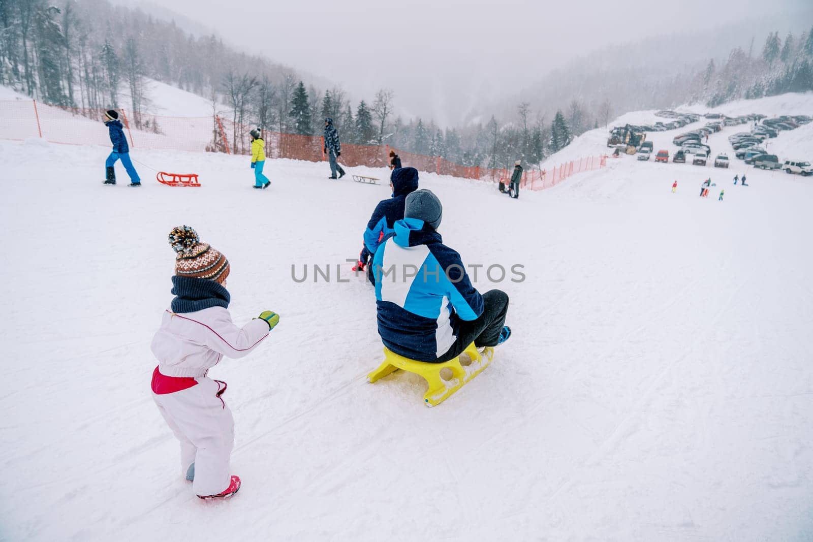 Small child pushes his father on a sled down a snowy hill and looks after him. Back view. High quality photo