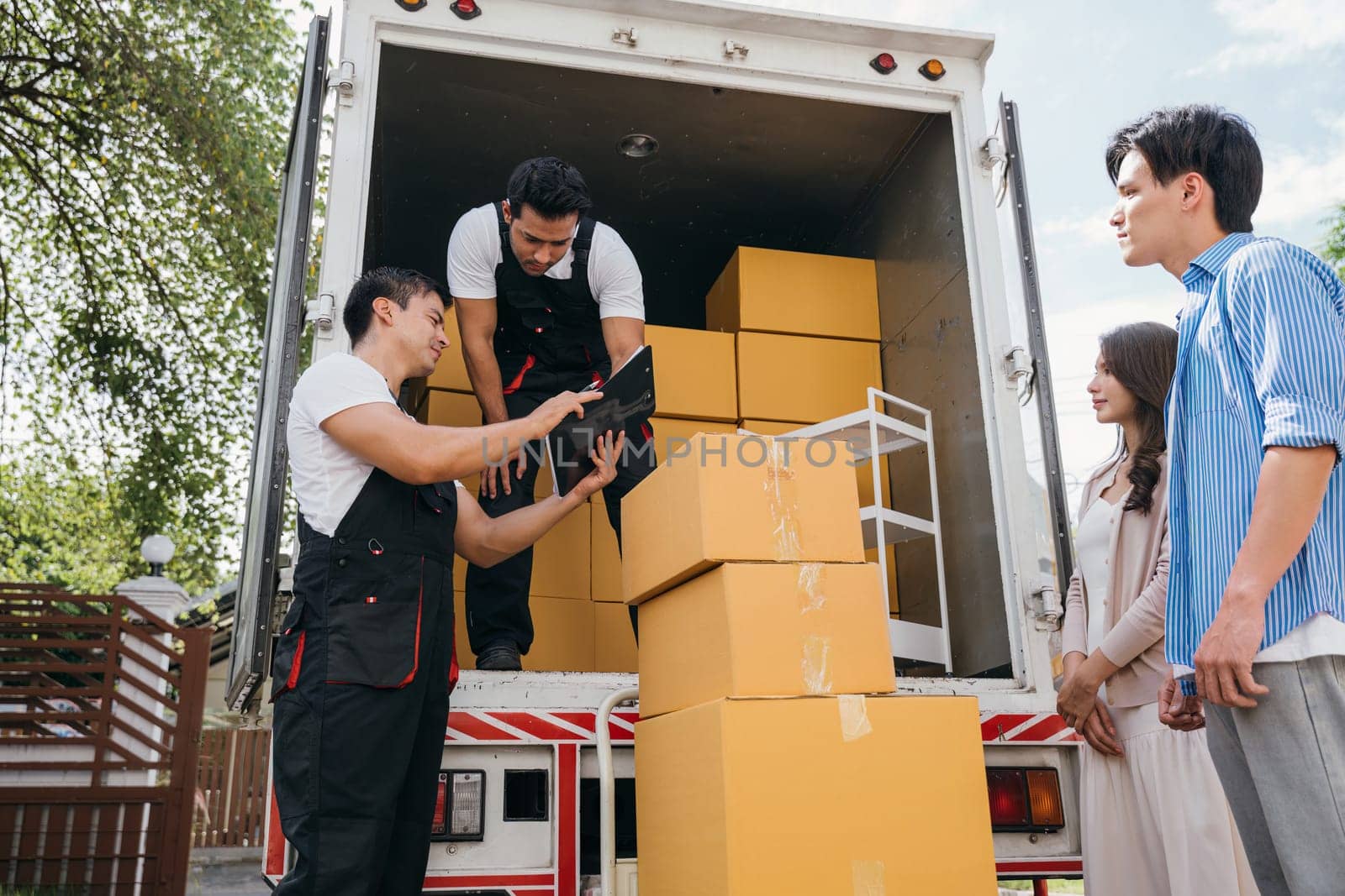 Newlywed couple satisfied signs delivery checklist with professional movers after furniture upload. Employees in uniform display teamwork for customer happiness. Moving Day Concept by Sorapop
