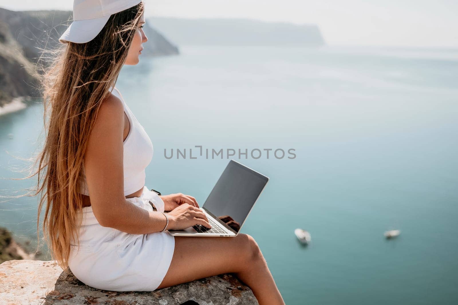 Successful business woman in yellow hat working on laptop by the sea. Pretty lady typing on computer at summer day outdoors. Freelance, travel and holidays concept.