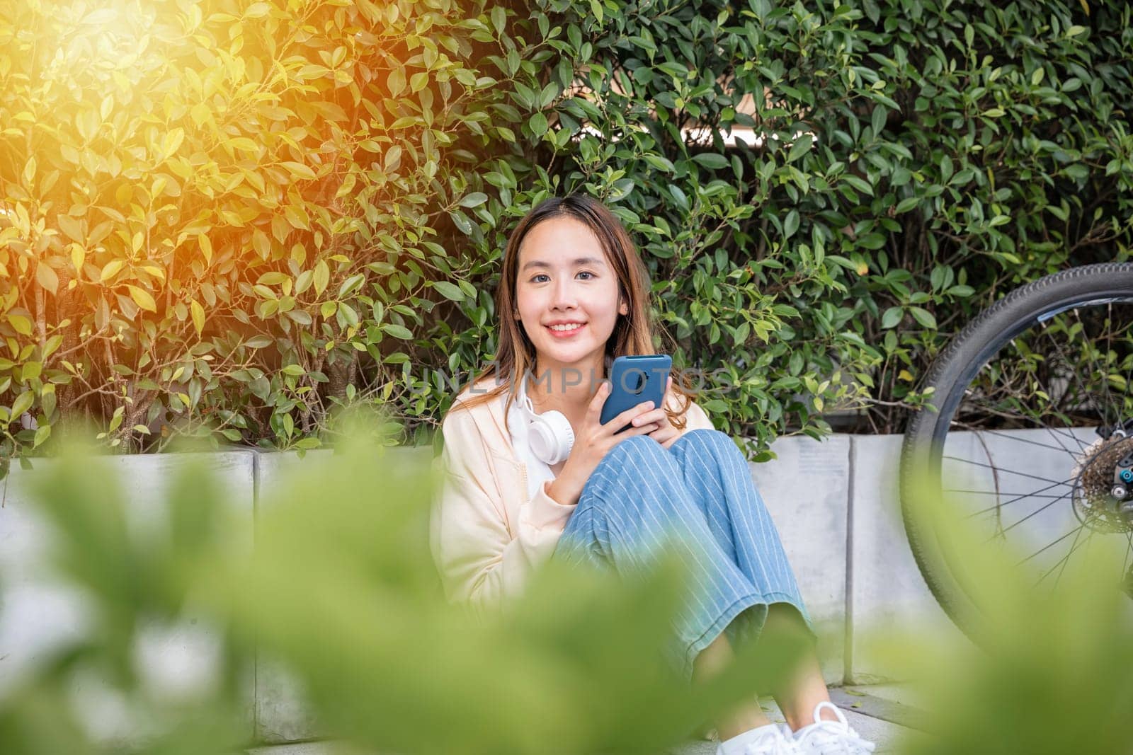 Beautiful spring day cute young woman sits in park with her bicycle listening to music through earphones and typing joyful message on her mobile phone. joyful expression reflects season happiness. by Sorapop