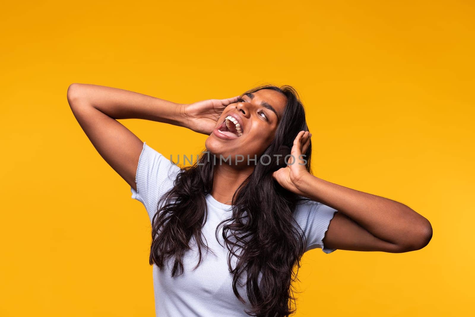 Happy young Indian woman wearing wireless headphones singing song out loud. Isolated on yellow background. Studio shot. by Hoverstock