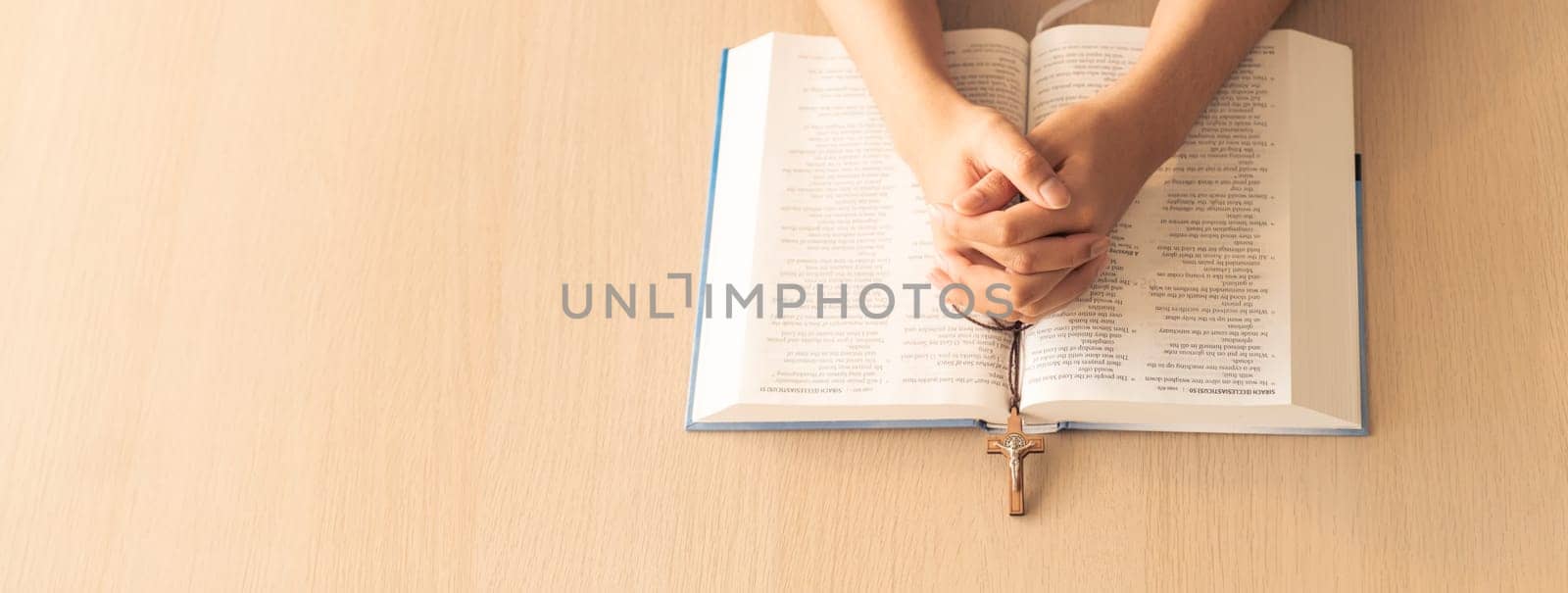 Cropped image of praying male hand holding cross on holy bible book at wooden table. Top view. Concept of hope, religion, faith, christianity and god blessing. Warm and brown background Burgeoning.