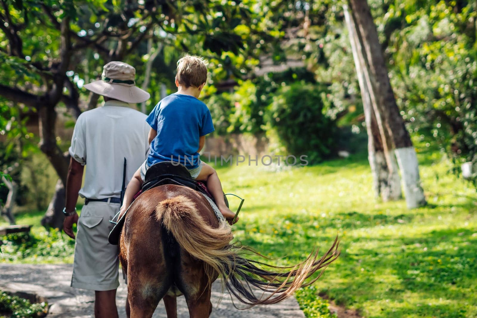 Blond boy riding horse in hotel park on the beach. Sunny summer day happy childhood.
