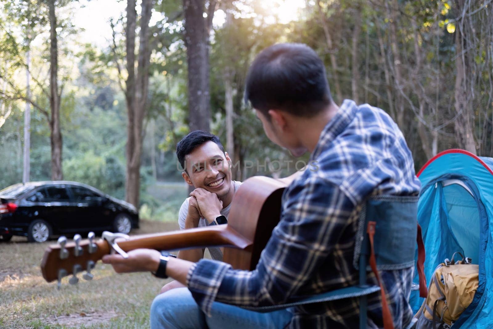 Asian LGBTQ couple enjoying nature, camping with tents in the forest area by the river, playing guitar. by wichayada