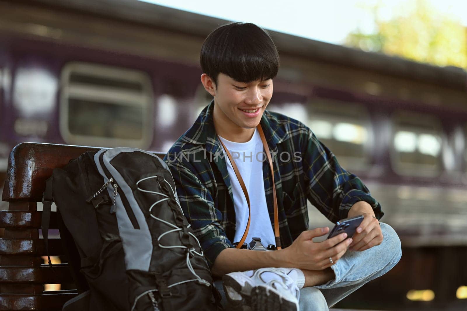 Smiling Asian male sitting on bench waiting for train and using mobile phone. Travel and lifestyle concept.