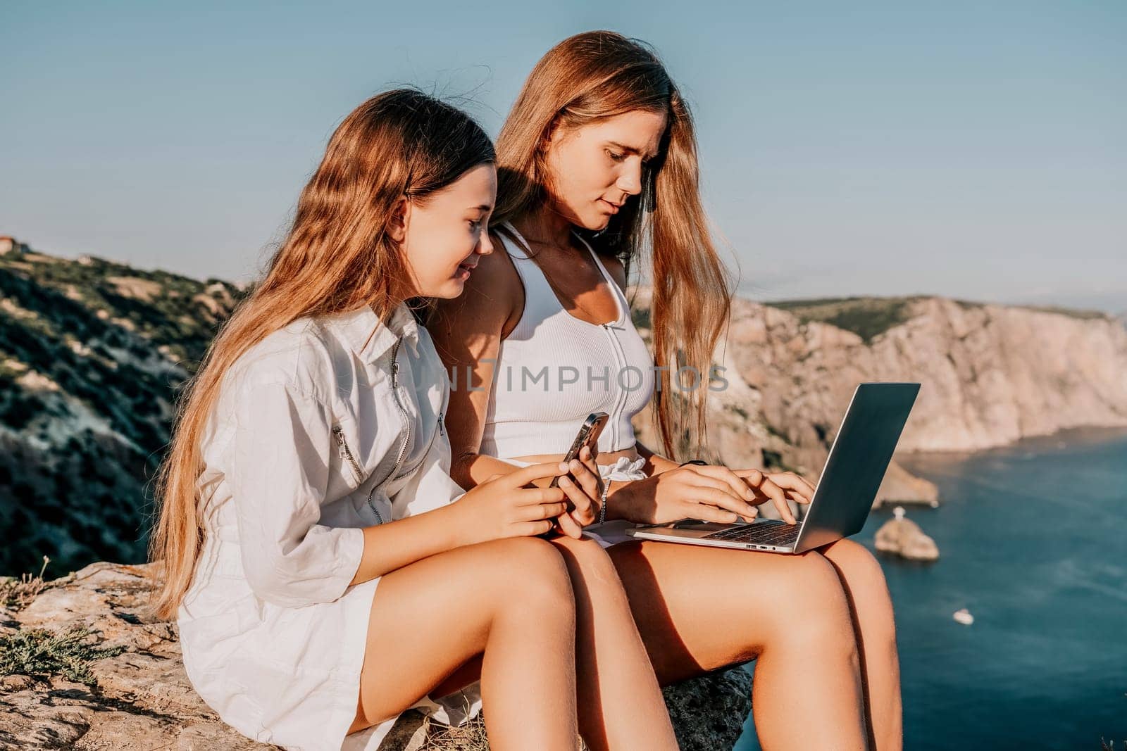 Digital nomad, Business woman working on laptop by the sea with daughter. Pretty lady typing on computer by the sea at sunset, makes a business transaction online. Freelance, remote work on vacation by panophotograph