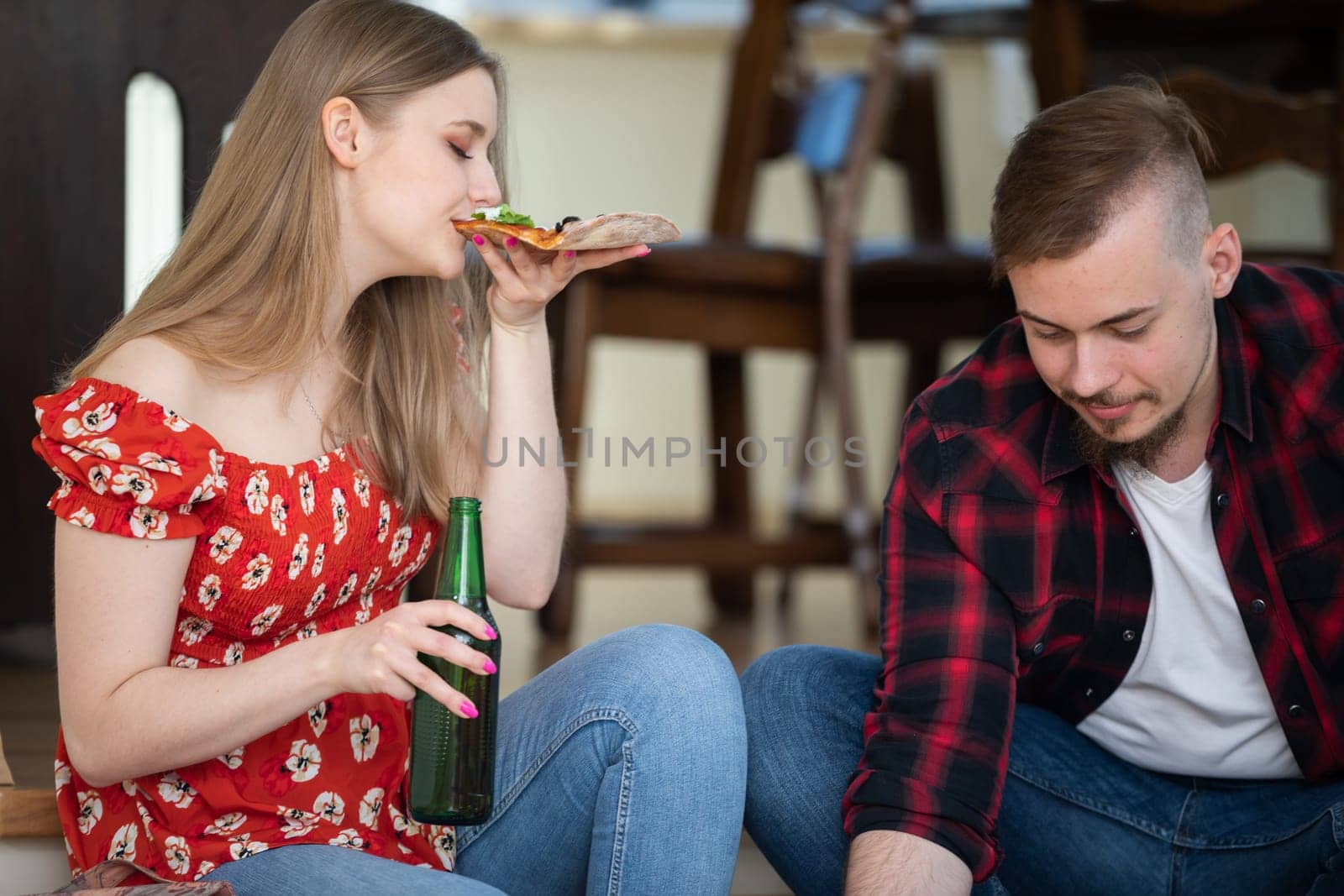 A hungry young girl bites into a slice of pizza with her eyes closed. She holds a bottle of beer in her other hand. Next to her sits a man with a beard and mustache.