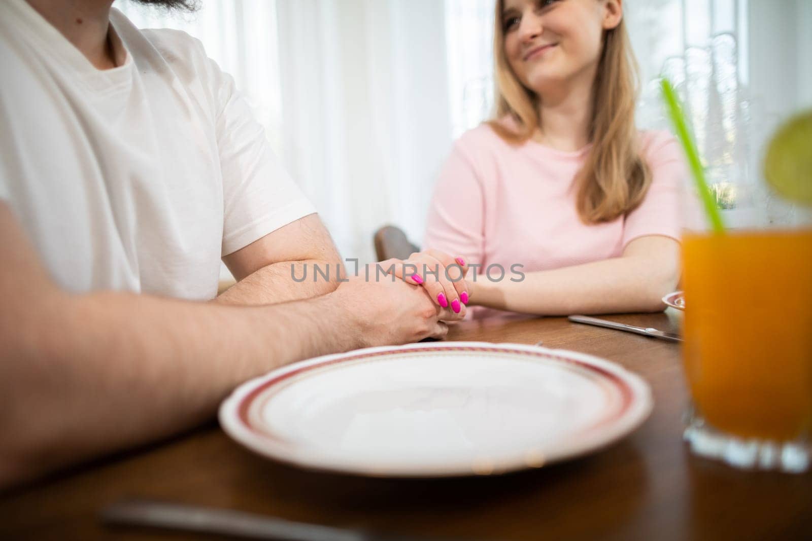 A man and a woman spend time together on a romantic date. They sit at a table and hold hands. A section of the table is visible in the foreground, with a plate and a glass of juice on it.