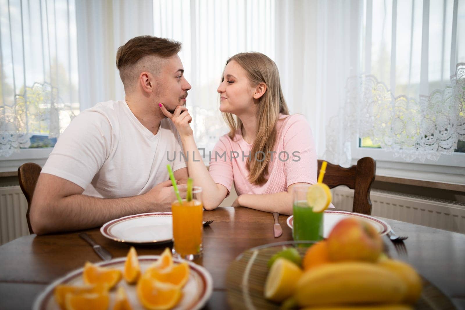 The woman holds her husband's chin and looks deeply into his eyes. The couple sits at a table set with fruit. In the background you can see a large window covered by a curtain.