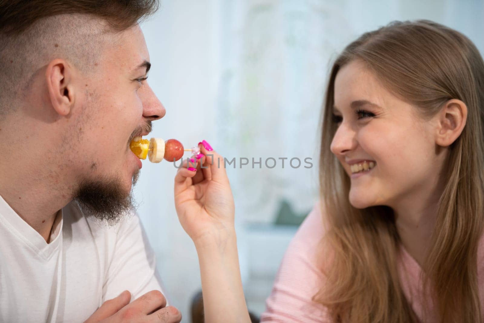 The girl feeds the man fruit. A grape, a slice of banana and an orange are impaled on a toothpick. The boy eats from his girlfriend's hand.