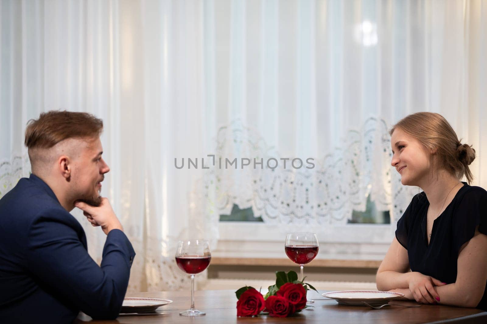 A woman and a man are sitting at a table facing each other. They both look at each other. There is a bouquet of red roses and two glasses of red wine on the table. In the background you can see a window covered with a curtain.