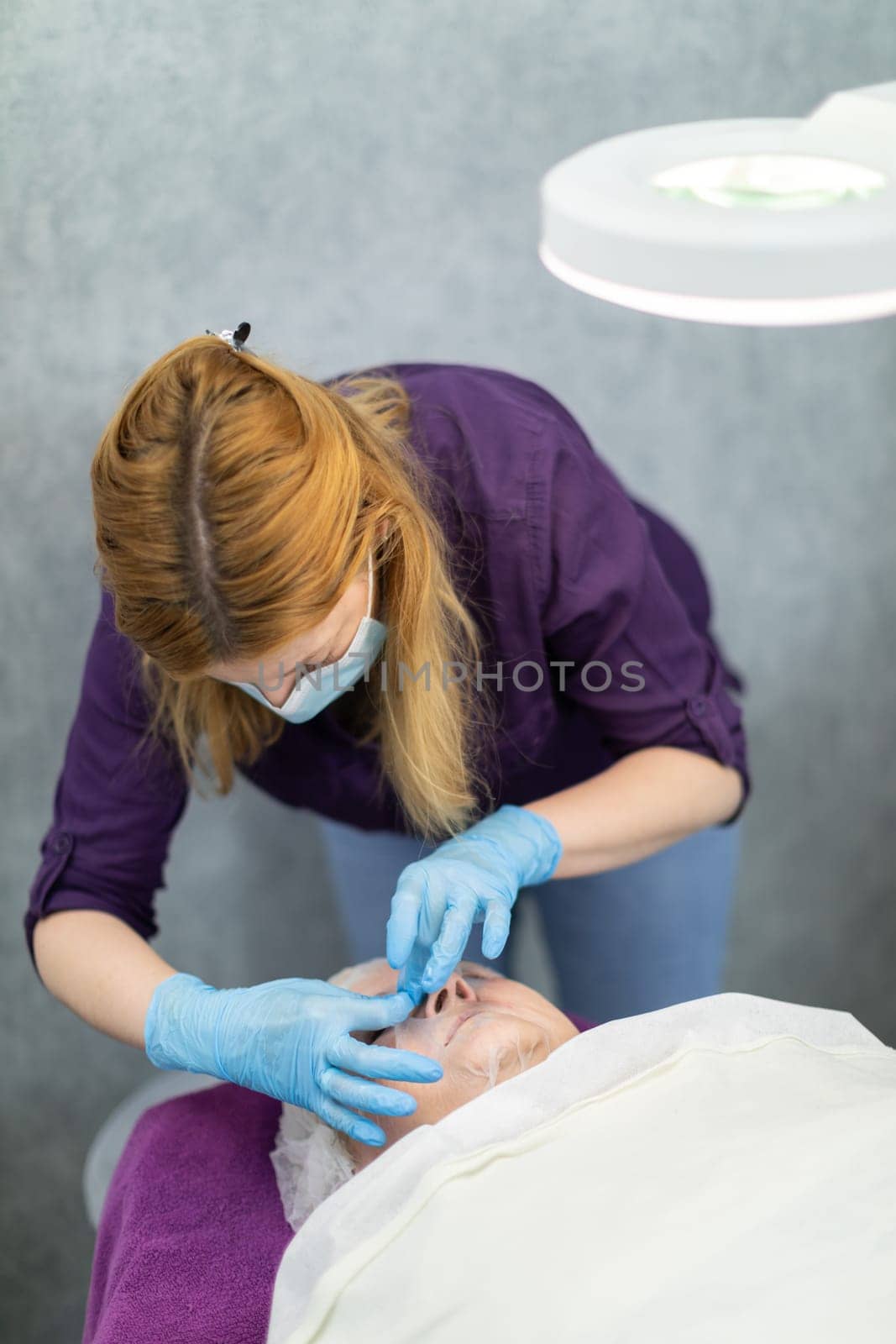 A young woman works in a beauty salon as a cosmetologist. She attends to her client, who is lying next to her on the cosmetic bed.