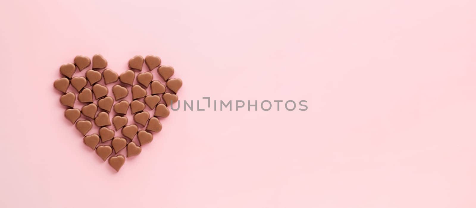 Heart made of romantic chocolate confections in heart shapes on pink background. Long horizontal banner with chocolate heart of milk chocolate sweet, copyspace. Topview flatlay. Valentines Day concept