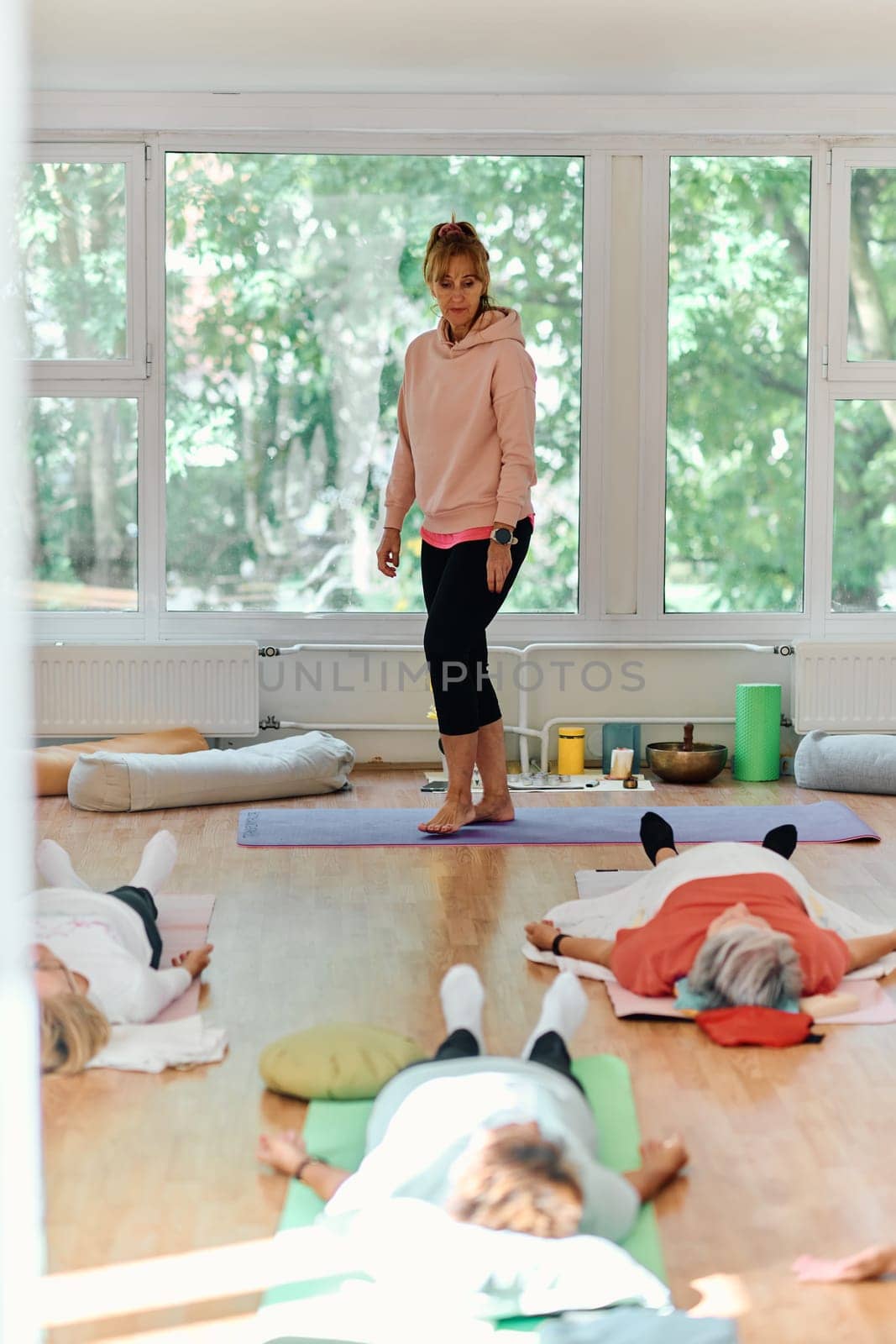 A skilled trainer oversees a group of senior women practicing various yoga exercises, including neck, back, and leg stretches, in a sunlit space, promoting wellness and harmony.