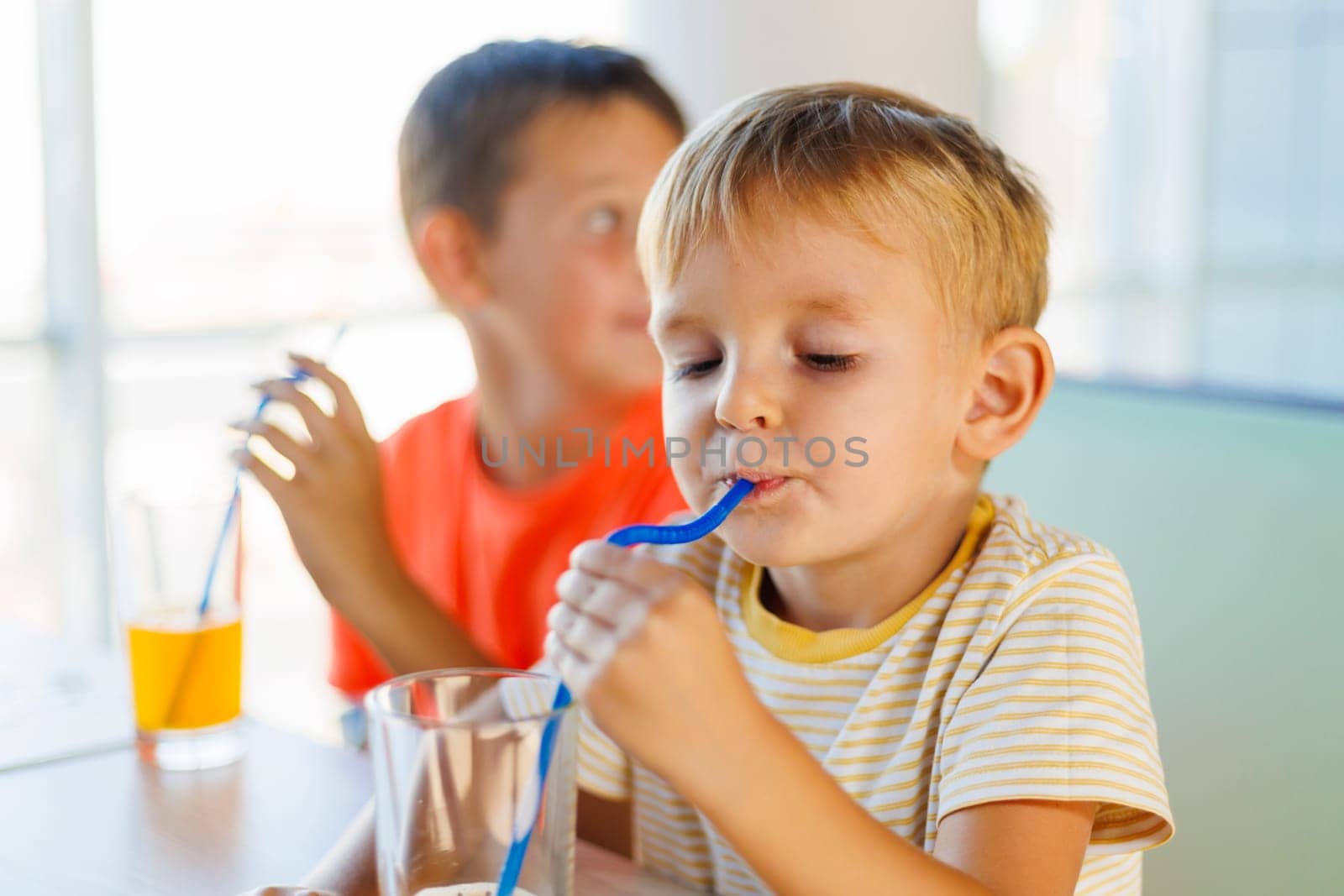 A young boy sipping juice through a straw with his brother in the background