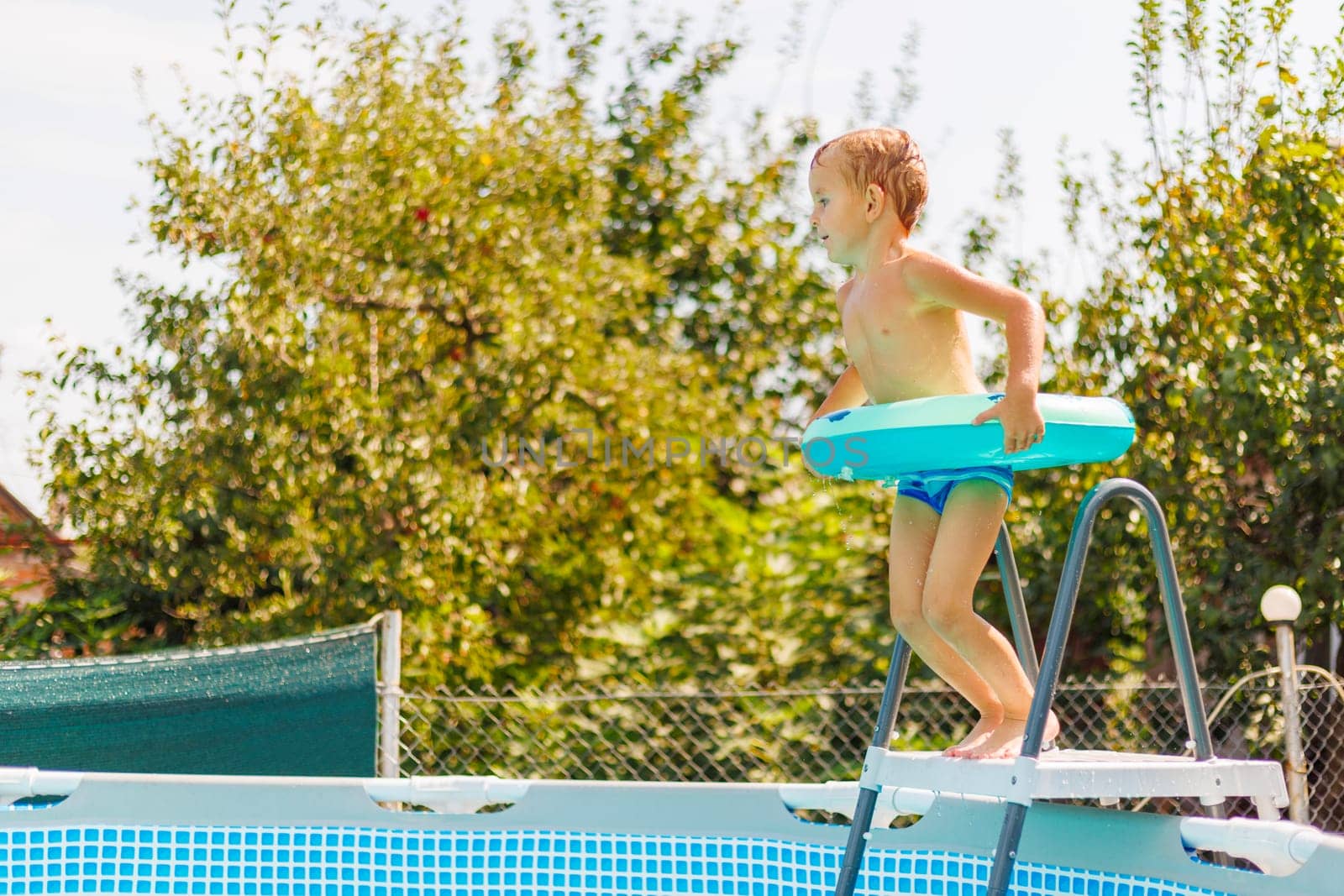 Excited young boy with a blue surfboard float preparing to jump into an above-ground pool on a sunny summer day, surrounded by greenery.