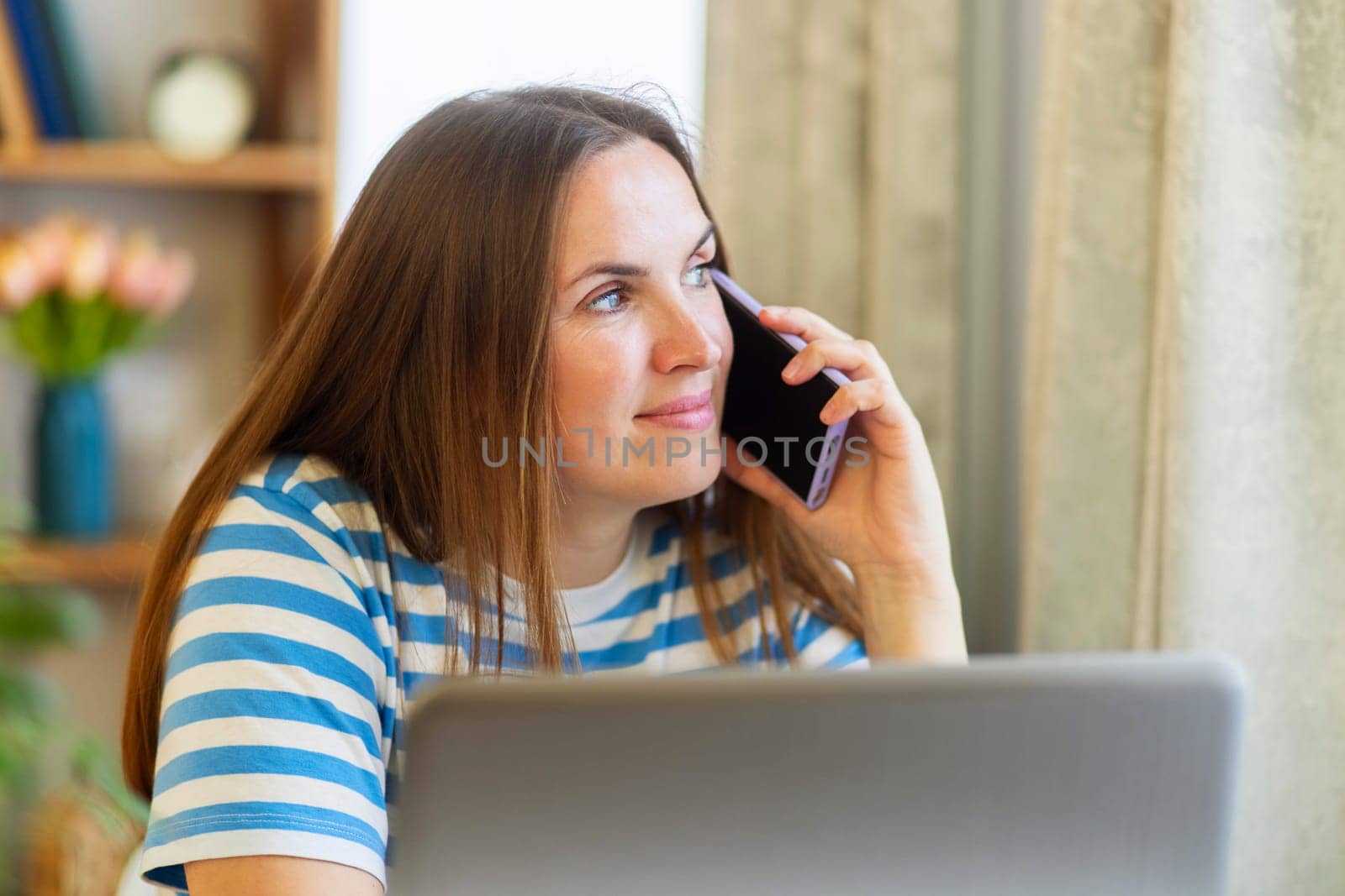 Smiling woman having a phone conversation while working on a laptop at home.