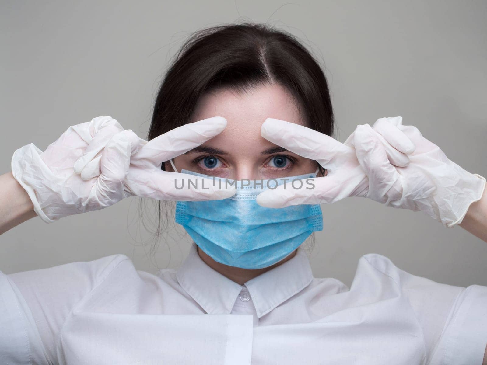 Young woman patient in a medical mask puts on protective surgical sterile gloves on her arm, on gray background, protection against coronovirus